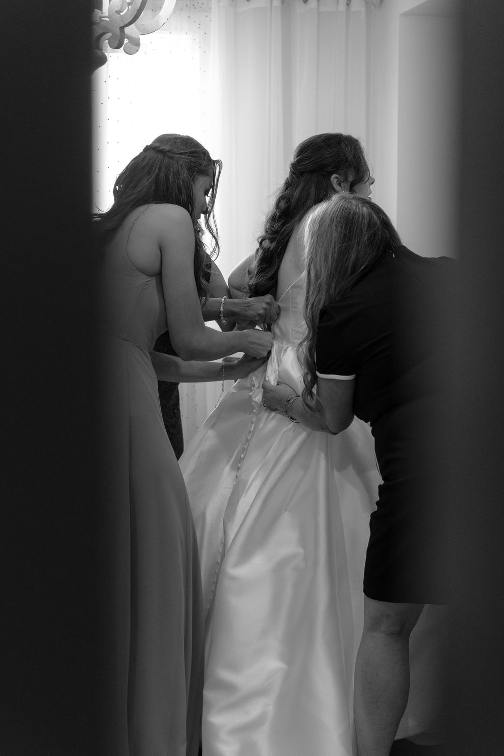 Bridesmaids helping bride into white wedding gown in black and white photo.