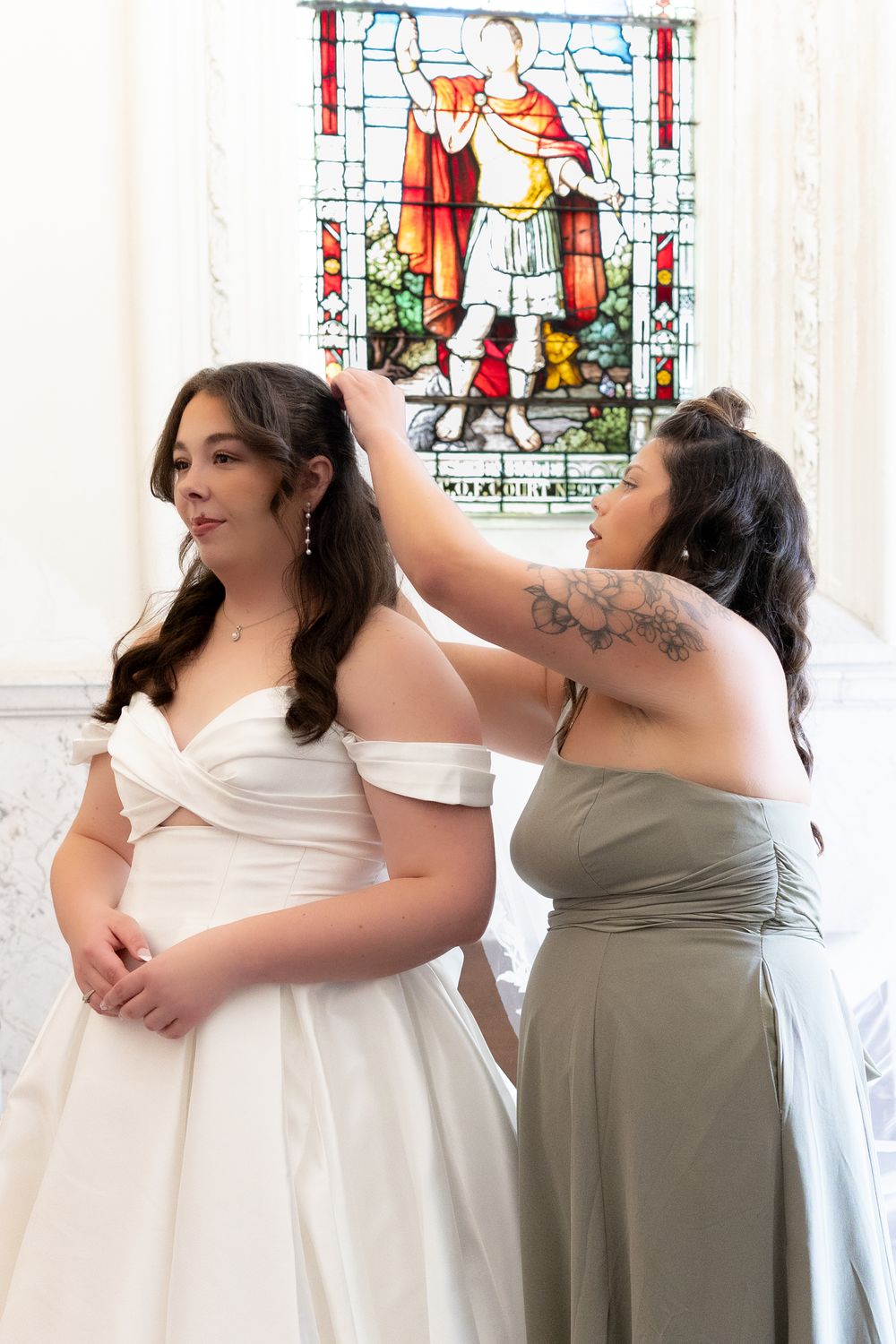 Bride in white off-shoulder gown gets hair styled by bridesmaid in sage dress before stained glass window.