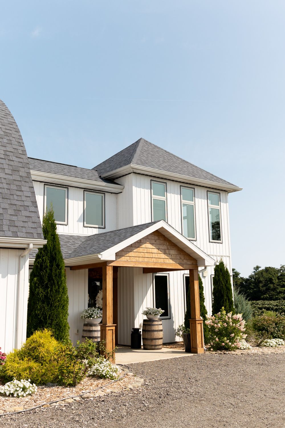 White two-story farmhouse winery entrance with wood-framed portico, barrel planters, cypress trees, and manicured gardens under blue sky.