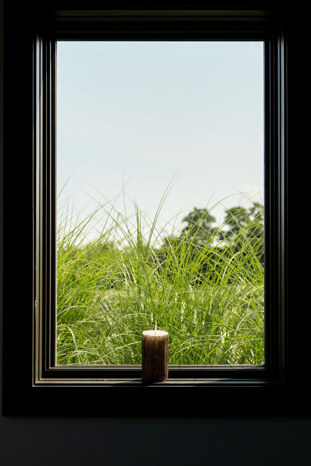 A brown pillar candle sits on a dark windowsill overlooking lush green grass and trees under a clear blue sky.