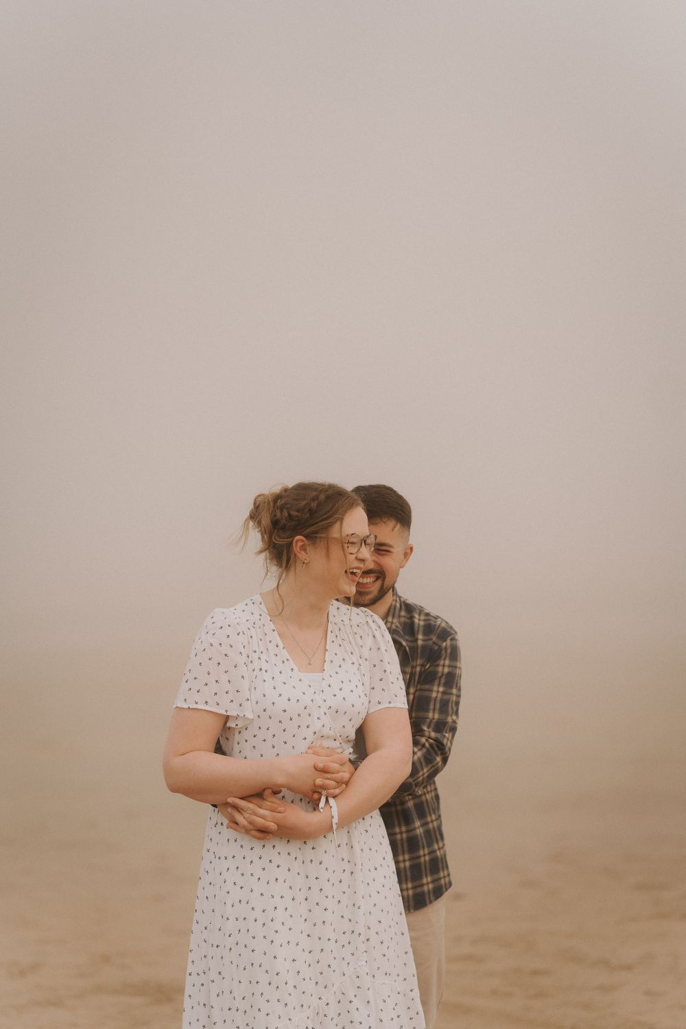 Couple laughing together on foggy beach, woman in white polka dot dress, man in plaid shirt.