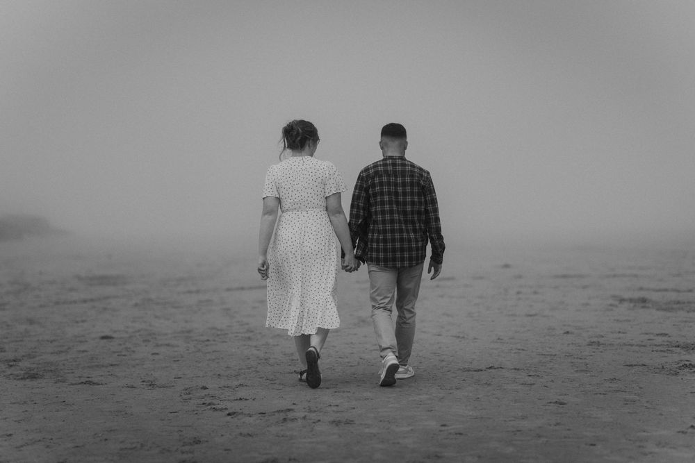 A couple walks hand in hand through misty fog on a beach, woman in white dress, man in plaid shirt.