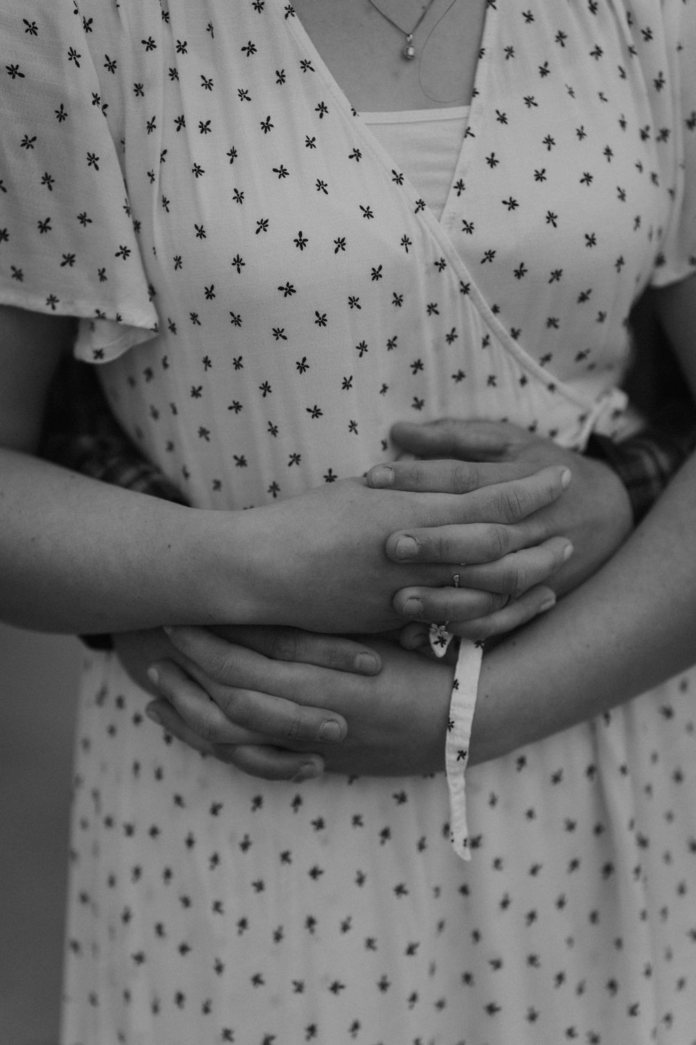 Black and white photo of a pregnant woman in a floral dress cradling her baby bump with both hands.