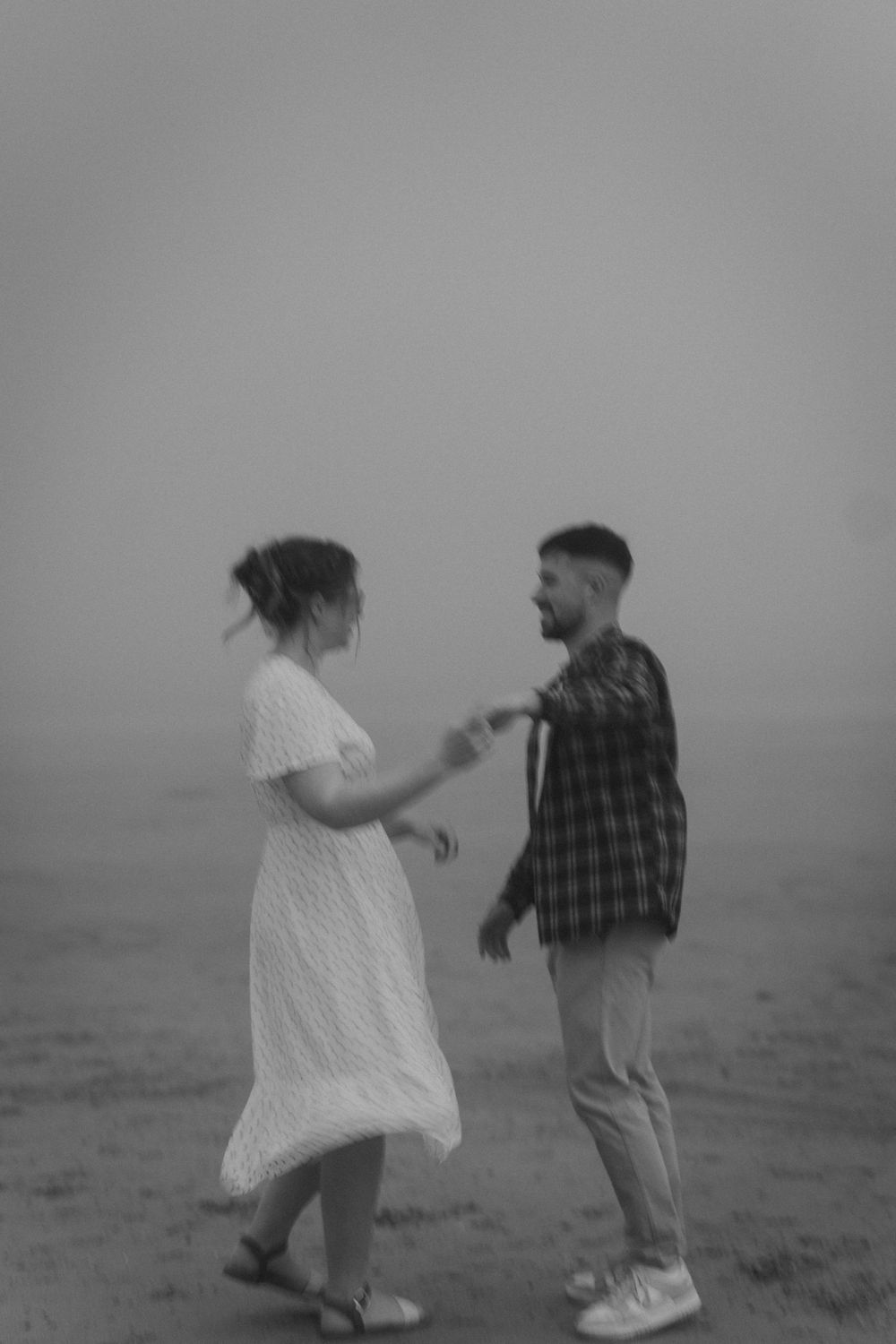 Black and white photo of joyful couple dancing on misty beach, woman in white dress, man in plaid.