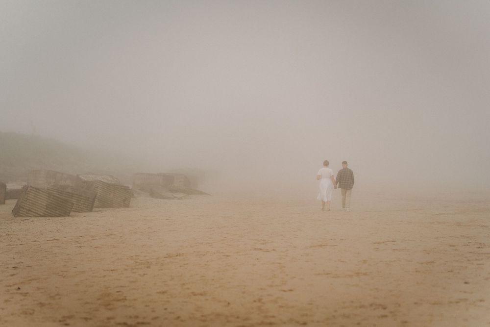 Two people walking on a foggy sandy beach with rocky outcrops disappearing into the mist.