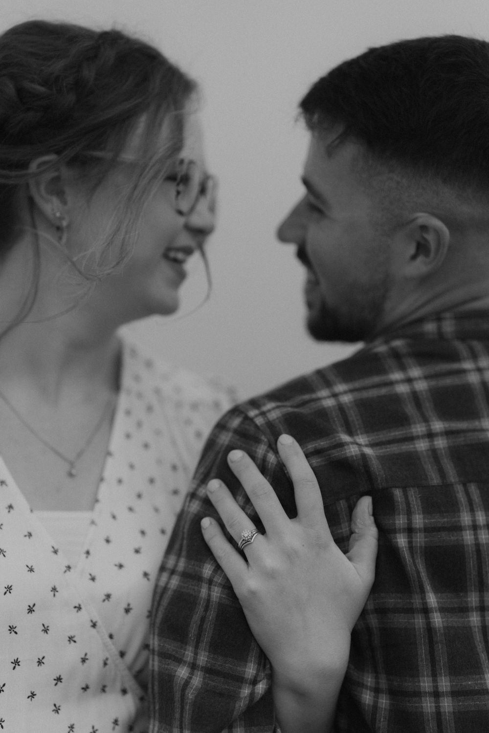 Black and white photo of smiling couple gazing at each other, woman showing engagement ring on mans shoulder.