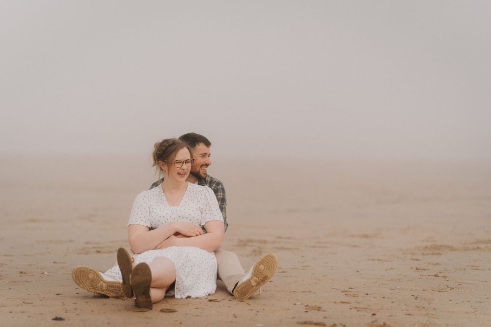 Couple sitting together on a sandy beach in foggy weather, woman in white dress, man embracing her from behind.