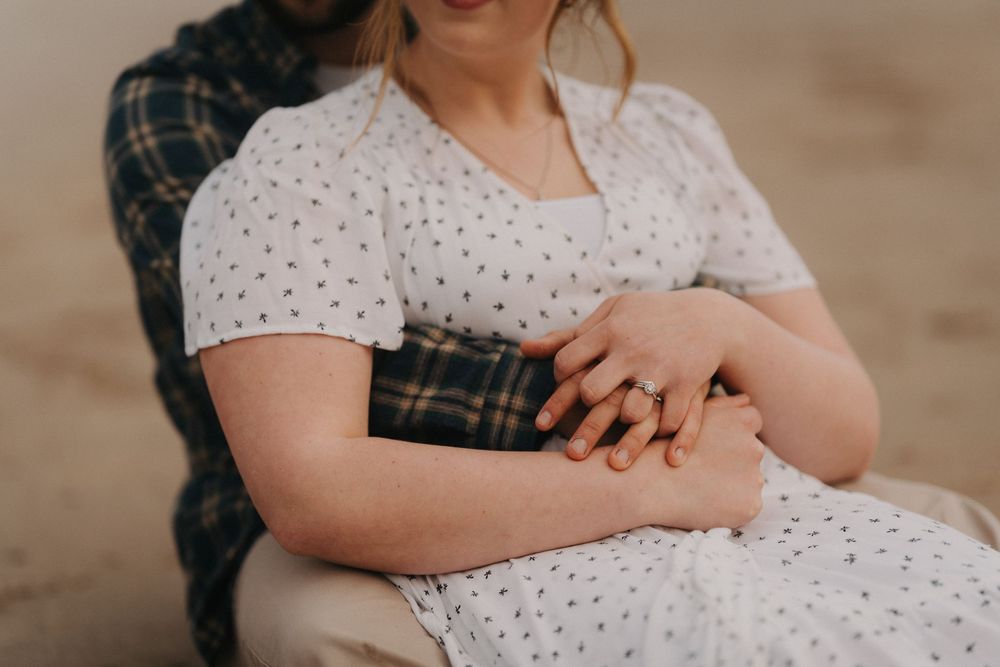 Close-up of couple holding hands, woman in white polka dot dress, man in plaid shirt on beach.