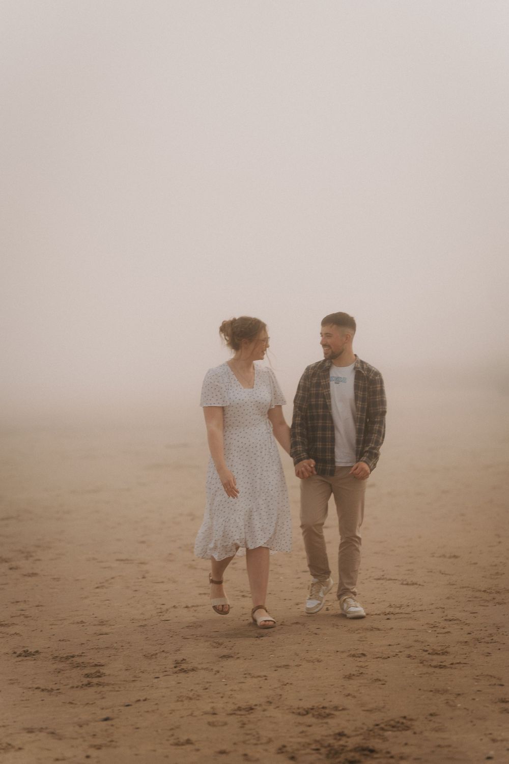 Couple walking hand in hand on a misty sandy beach, woman in white dress and man in plaid shirt.