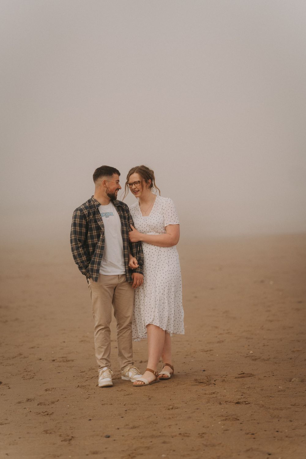 A couple shares a romantic moment on a foggy beach, the woman in a white dress and the man in a plaid shirt.