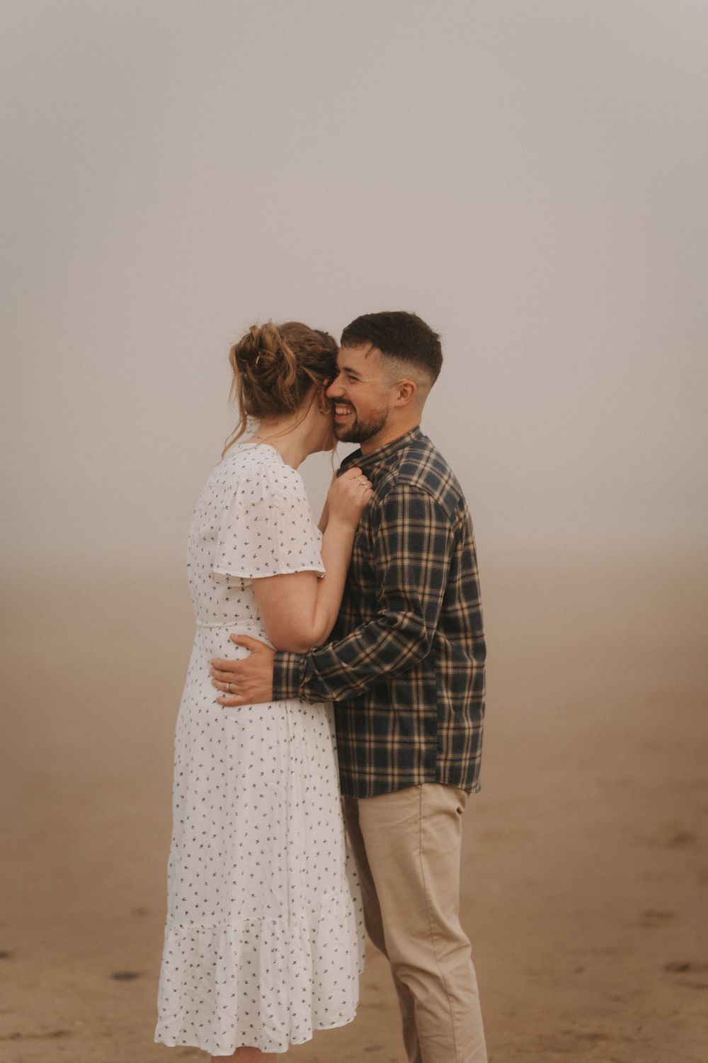 Couple embracing on a misty beach, woman in white polka dot dress and man in plaid shirt smiling.