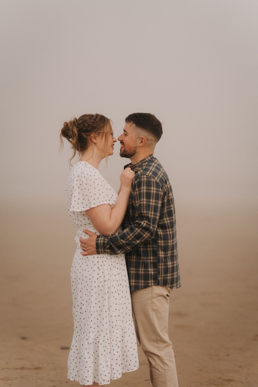 Couple sharing a romantic almost-kiss embrace on a misty beach, woman in white floral dress, man in plaid shirt.