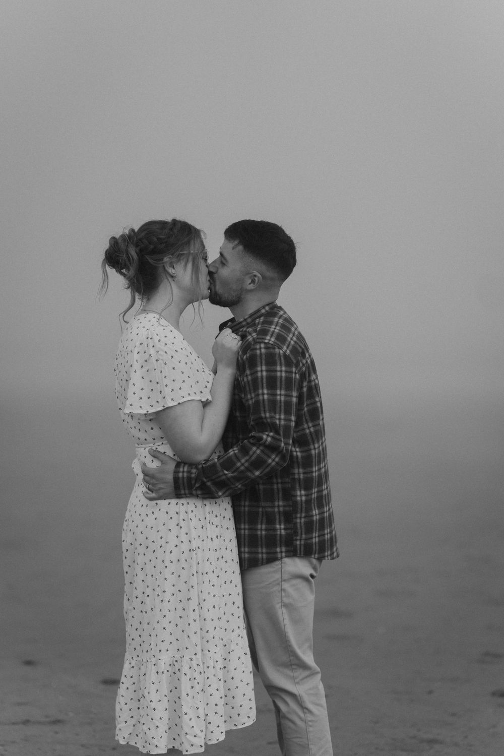 Black and white photo of a couple kissing in the fog, woman in polka dot dress embraced by man in plaid shirt.