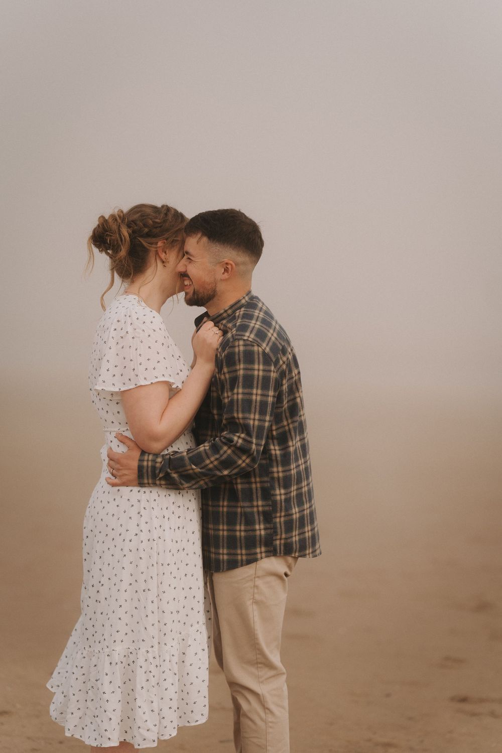 Romantic couple nearly kissing on foggy beach, woman in white dress and man in plaid shirt embracing.