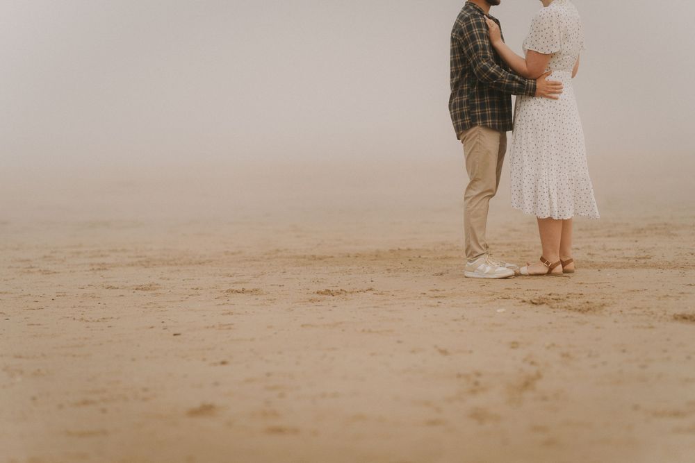 Couple embracing on a misty sandy beach, man in plaid shirt and khakis, woman in white dress.