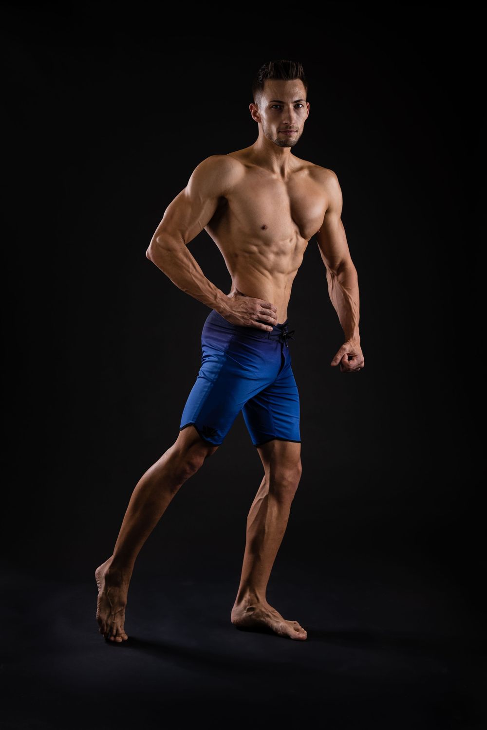 Muscular shirtless man in blue compression shorts posing confidently against a dark black background.
