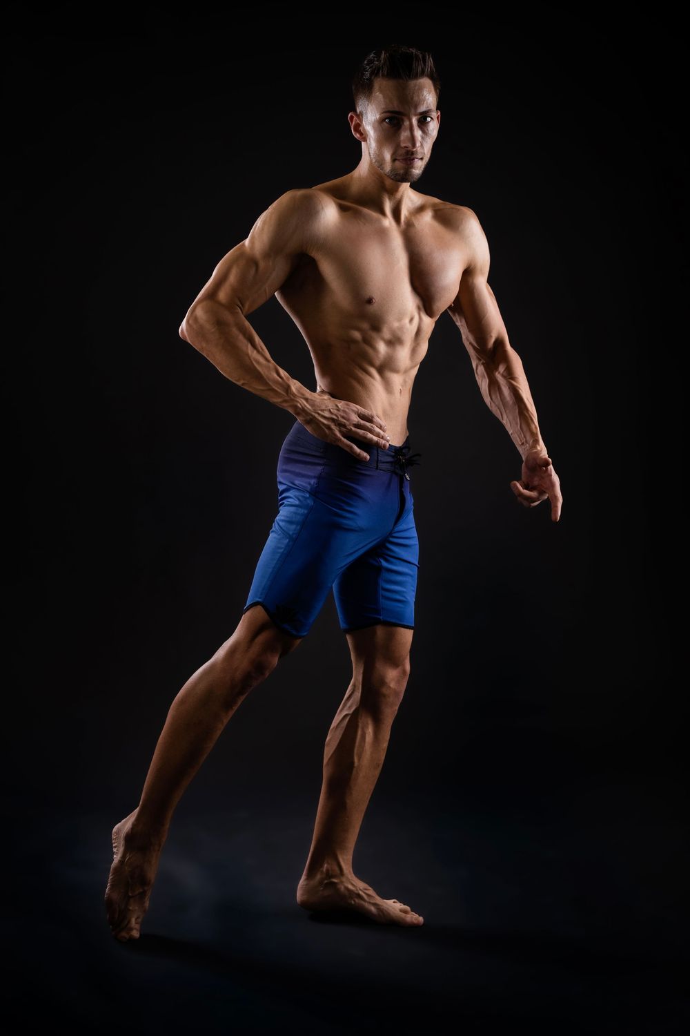 Muscular shirtless man in blue shorts posing against dark background, showcasing defined abs and athletic physique.