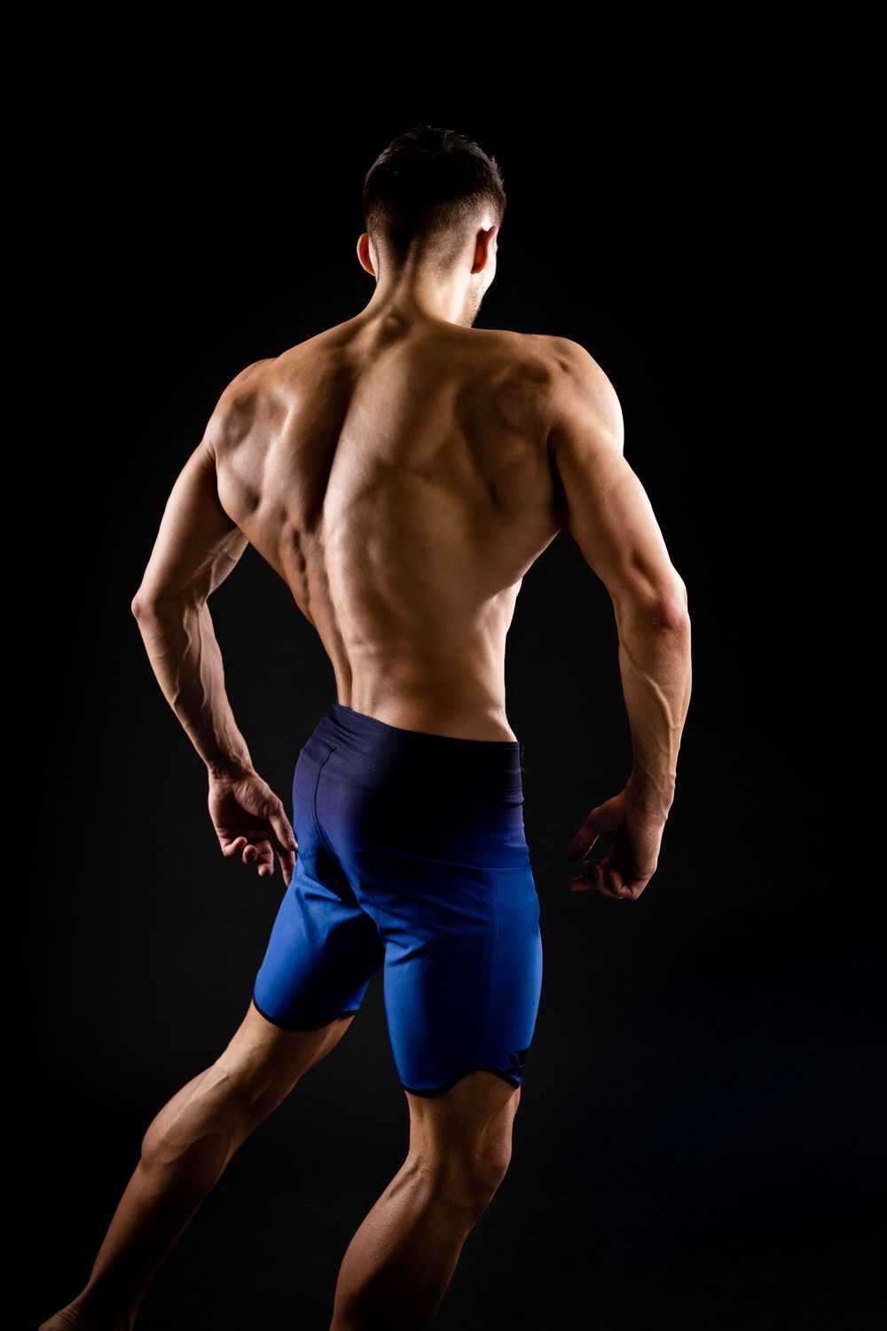 Muscular man in blue shorts showing defined back muscles against a dark background.