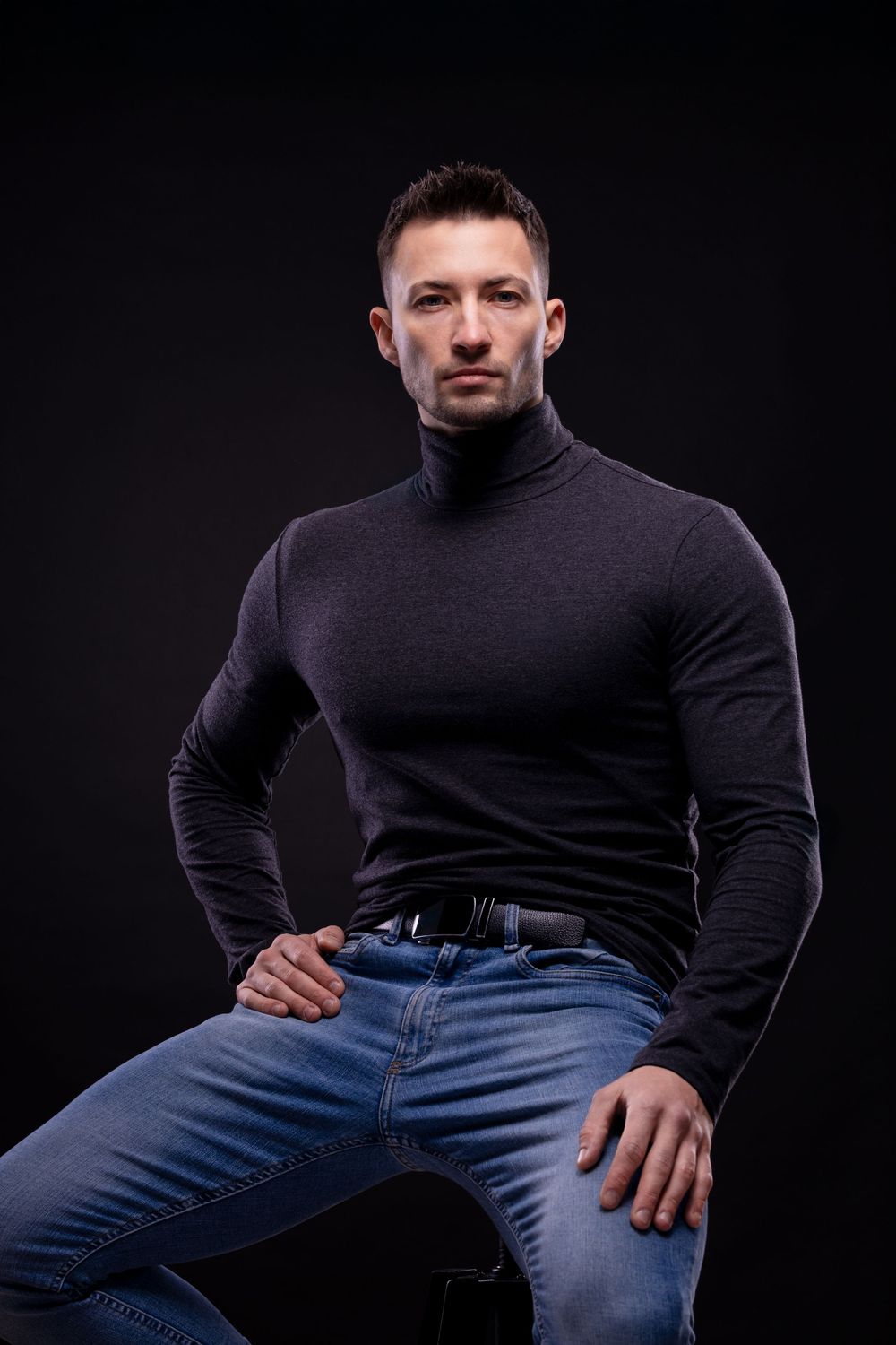 Muscular man in black turtleneck and blue jeans posing confidently against a dark studio background.