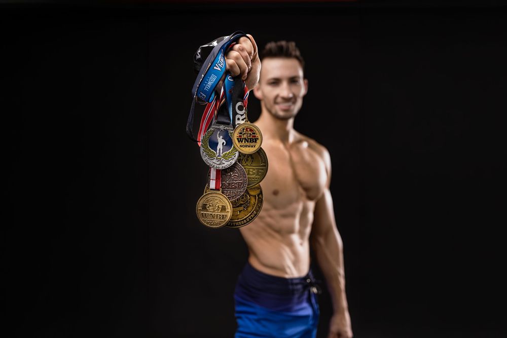 Shirtless male athlete proudly displaying multiple gold medals and trophies on dark background.