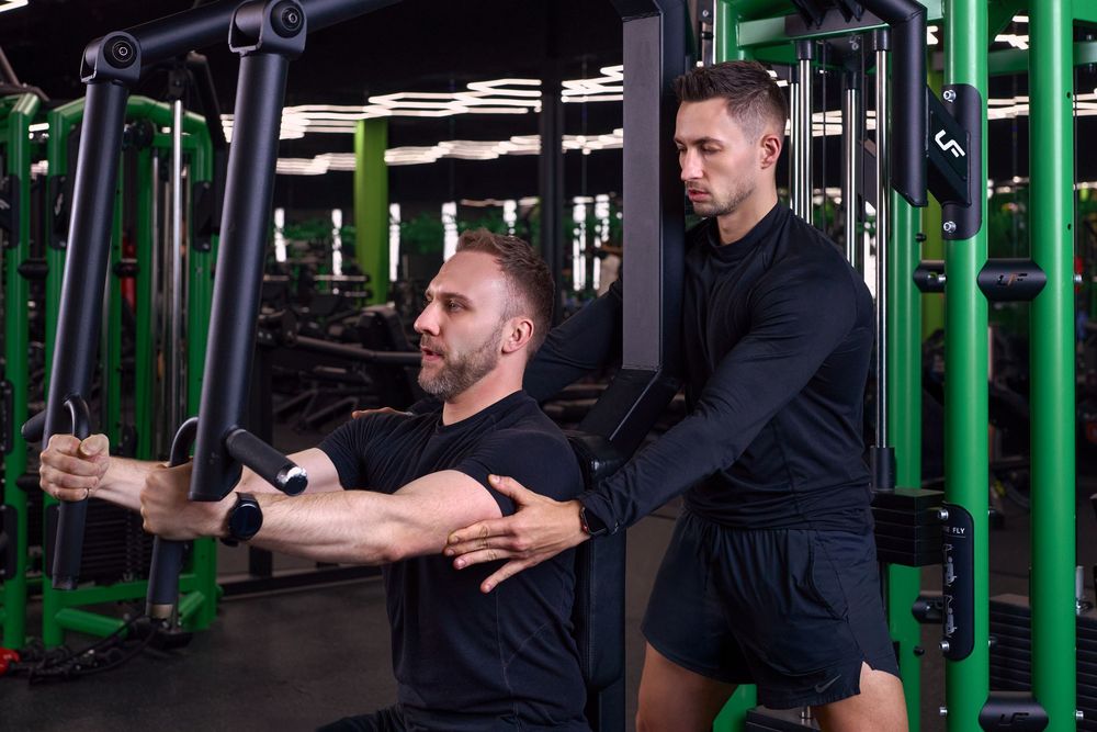 A personal trainer assists a man performing cable fly exercises on a gym machine, both wearing black athletic wear.