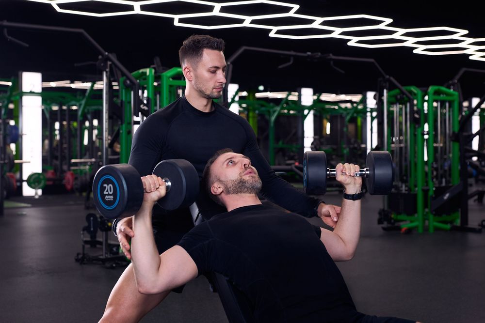 A personal trainer spots a man doing dumbbell bench press in a modern gym with hexagonal ceiling lights.