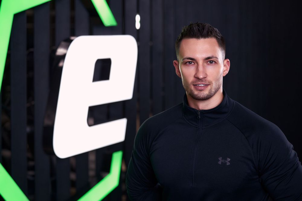 Man in black Under Armour jacket poses confidently in front of a green and black gym logo backdrop.