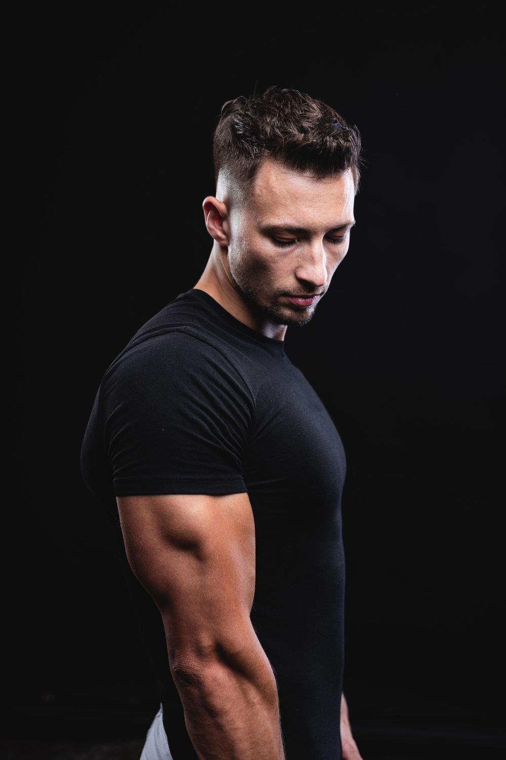 Muscular man in black t-shirt posing against dark background, showcasing athletic physique and defined arms.