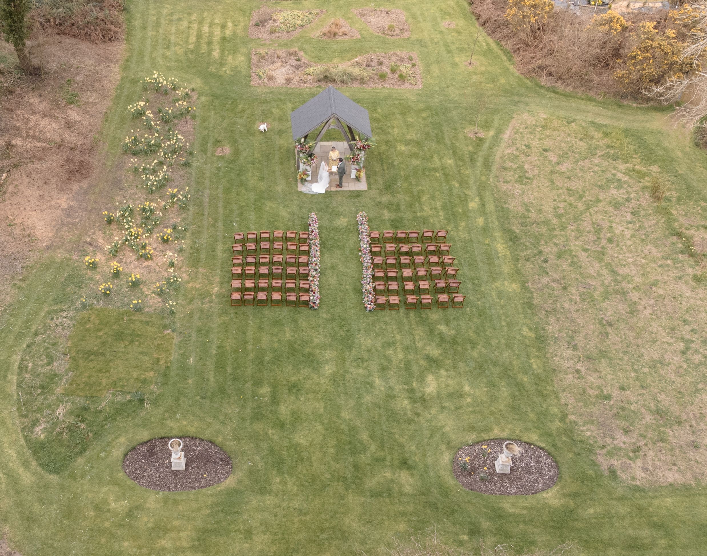 Aerial view of an outdoor wedding venue with rows of chairs facing a garden altar surrounded by manicured green lawns.