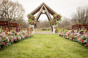 Outdoor wedding ceremony aisle lined with colorful flowers leading to a wooden arch adorned with pink and red floral arrangements.