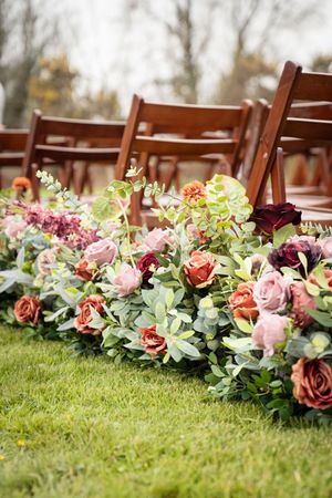 Lush floral aisle arrangement with pink, burgundy, and rust roses mixed with eucalyptus beside wooden chairs at outdoor wedding.