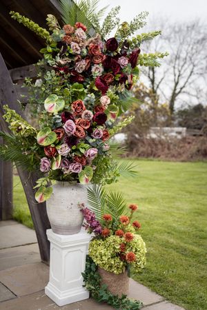 Lush floral arrangement with deep red roses and greenery in a stone vase on a white pedestal at an outdoor wedding venue.