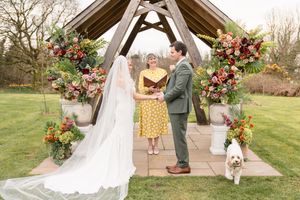 Bride and groom exchange vows outdoors under wooden arbor adorned with colorful floral arrangements, with officiant and white dog nearby.