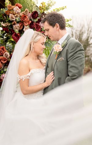 Bride and groom share an intimate moment in front of a floral arch with roses in autumn tones.