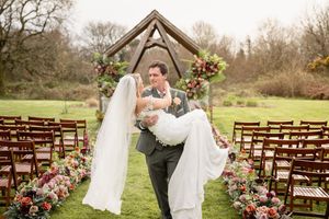 Groom carries bride in white gown and veil at outdoor wedding ceremony with floral arch and wooden chairs.