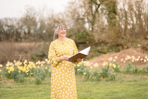 Woman in yellow polka dot dress holding open book standing in garden with daffodils blooming in background.