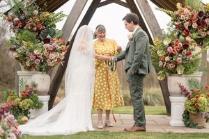 Bride and groom exchange vows under a rustic wooden arch adorned with lush autumn floral arrangements outdoors.