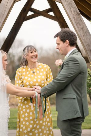 Officiant in yellow polka dot dress laughs joyfully during outdoor wedding hand-fasting ceremony under wooden arch.