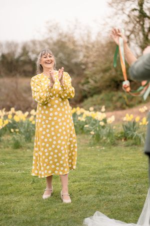 Smiling older woman in yellow polka dot dress clapping outdoors among daffodils at a wedding.