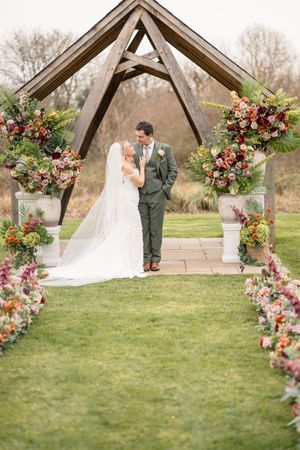 Bride and groom share a romantic moment under a wooden arch lined with vibrant autumn flowers and a floral aisle.