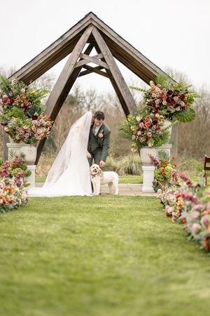 Bride and groom greet their fluffy dog at floral-adorned outdoor wedding altar with colorful blooms.