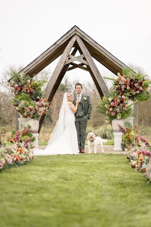 Smiling bride and groom pose with their white dog under a rustic wooden arch with vibrant floral arrangements.