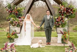 Bride and groom pose with their white dog under a rustic floral arch at an outdoor garden wedding ceremony.
