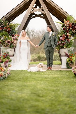 Bride and groom hold hands at floral wedding altar as their white dog rests on the grass aisle.
