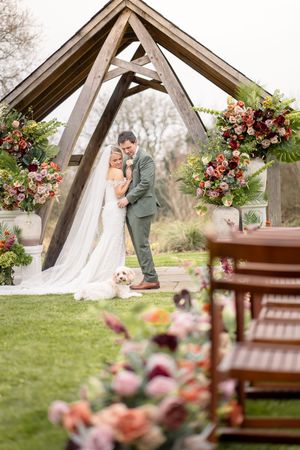 Bride and groom embrace under a rustic wooden arch adorned with colorful floral arrangements at an outdoor wedding ceremony.