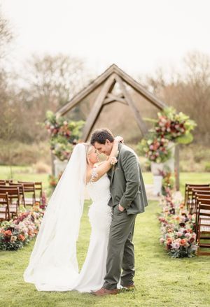 Bride and groom share a romantic kiss at outdoor wedding ceremony with floral arch and colorful flower arrangements.