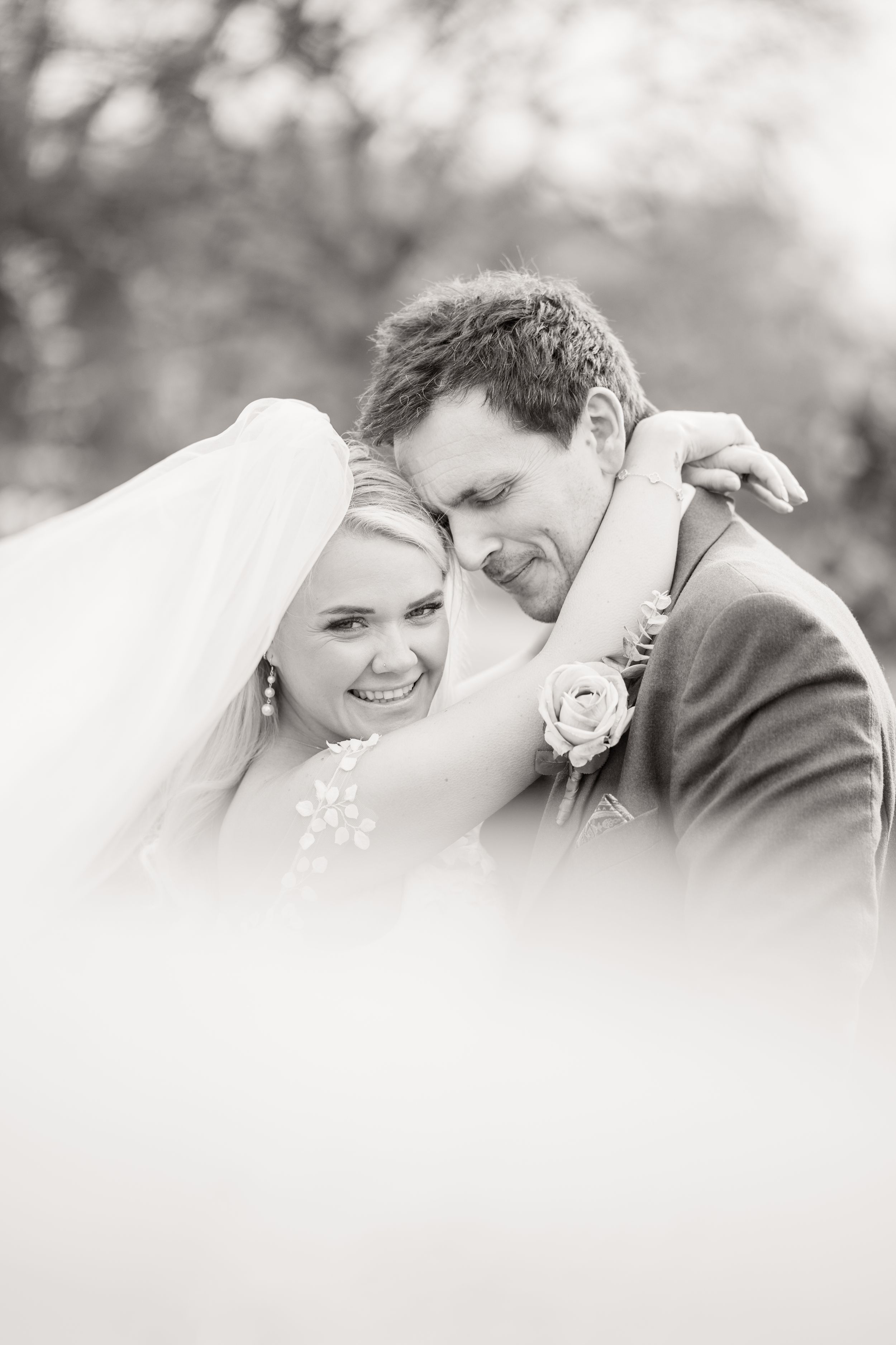 Black and white portrait of smiling bride embracing groom tenderly under her flowing wedding veil outdoors.
