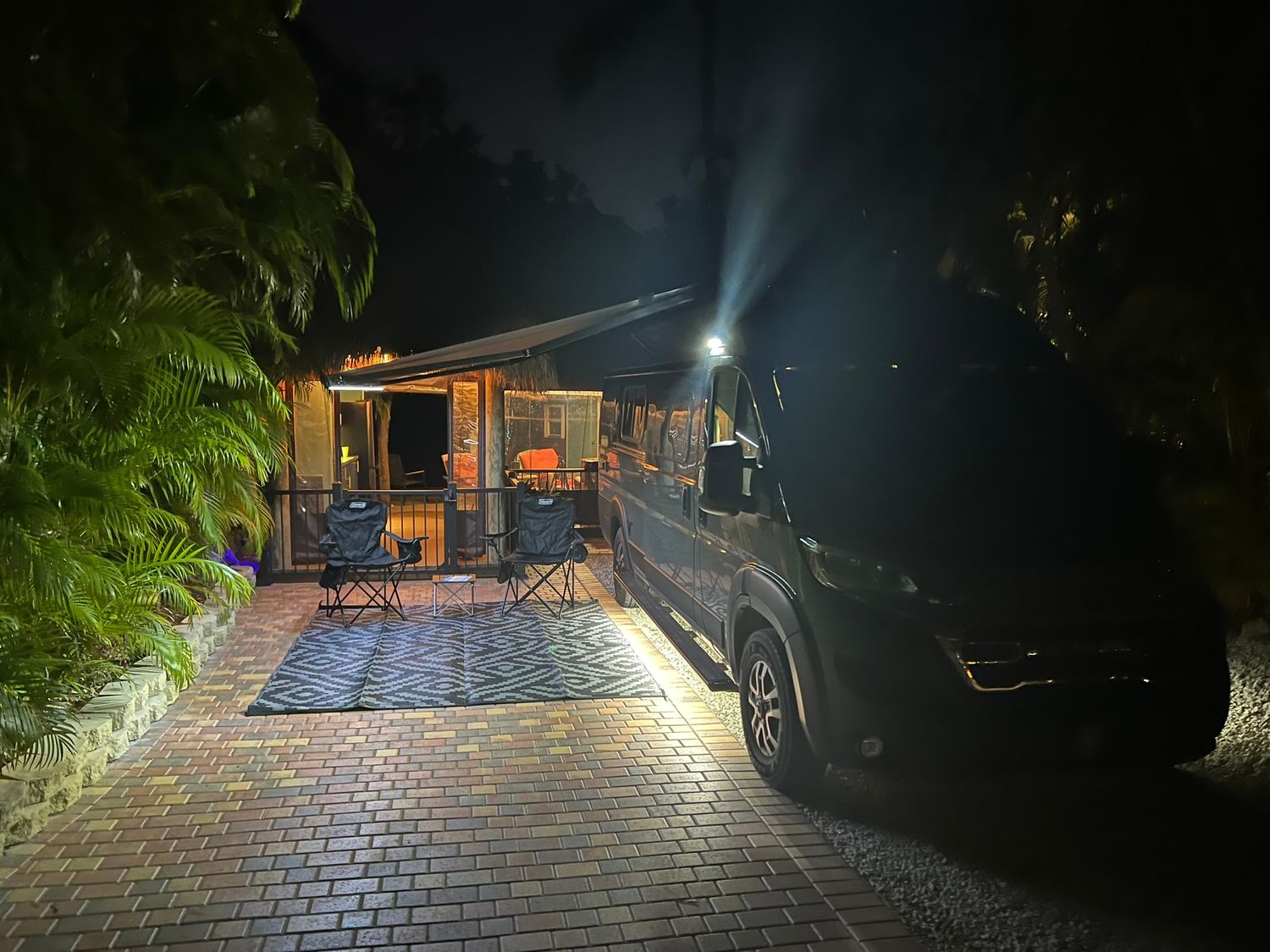 A motorhome parked on a brick driveway at night with outdoor chairs and warm lights glowing near tropical trees.