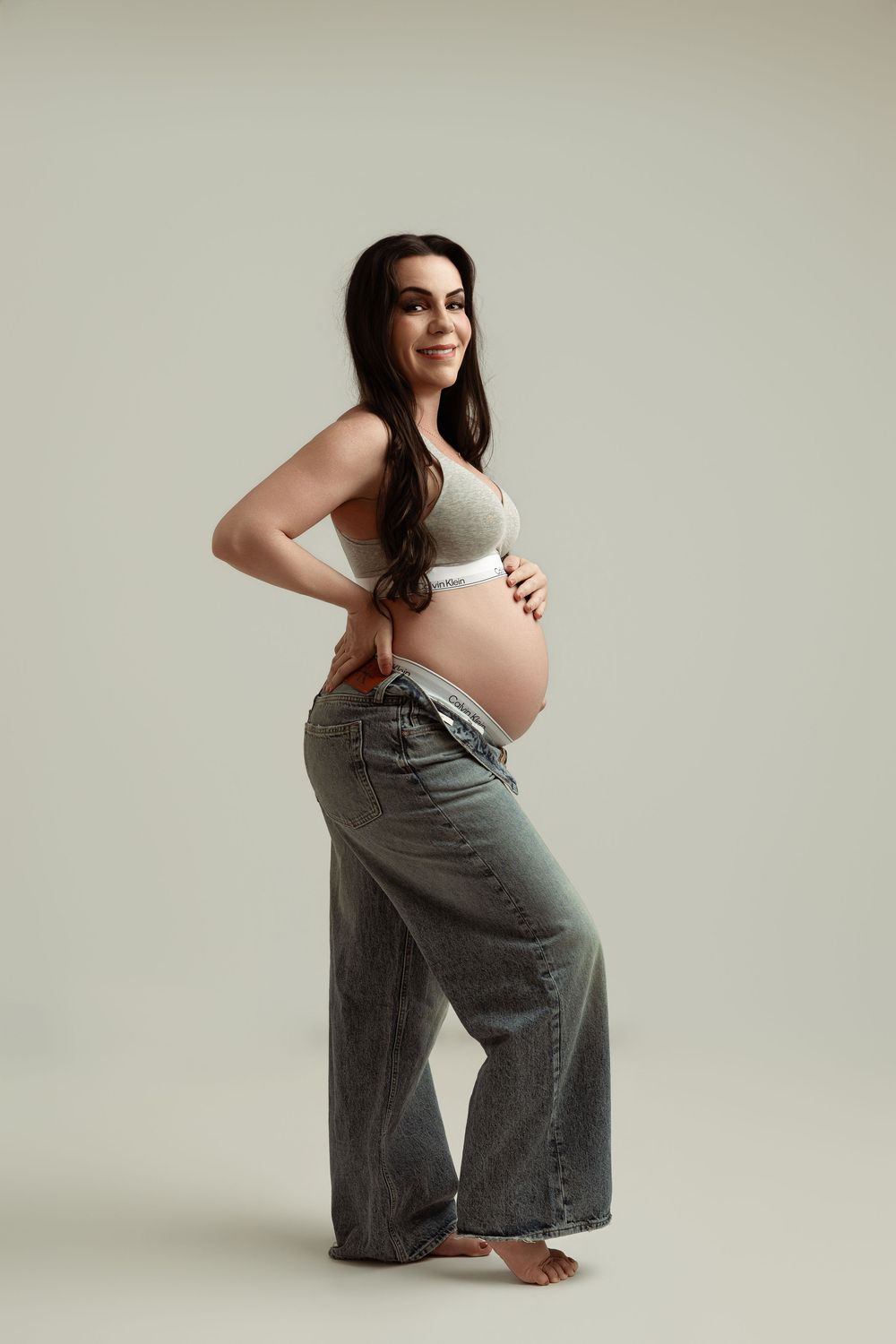Smiling pregnant woman in gray bralette and loose jeans cradling her baby bump against a neutral background.