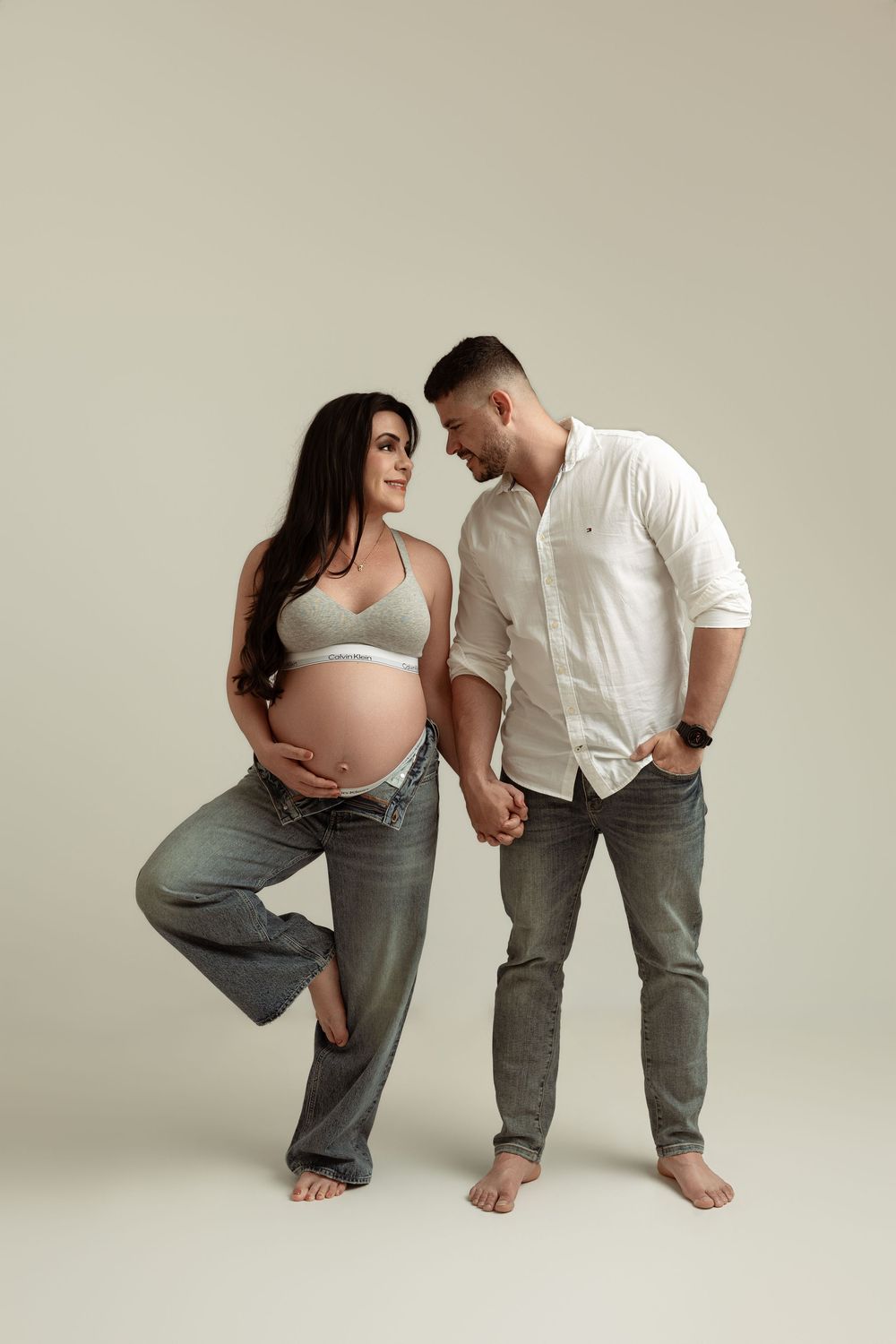 Pregnant couple holding hands in studio, woman in gray bra and jeans showing baby bump, man in white shirt.